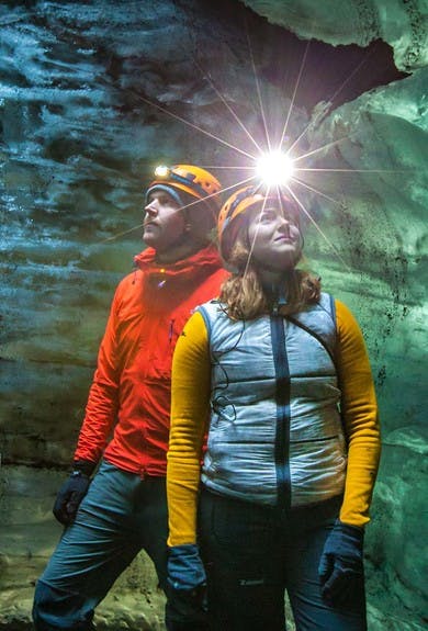 A couple standing in an ice cave in Iceland, wearing helmets with headlamps and gazing upwards