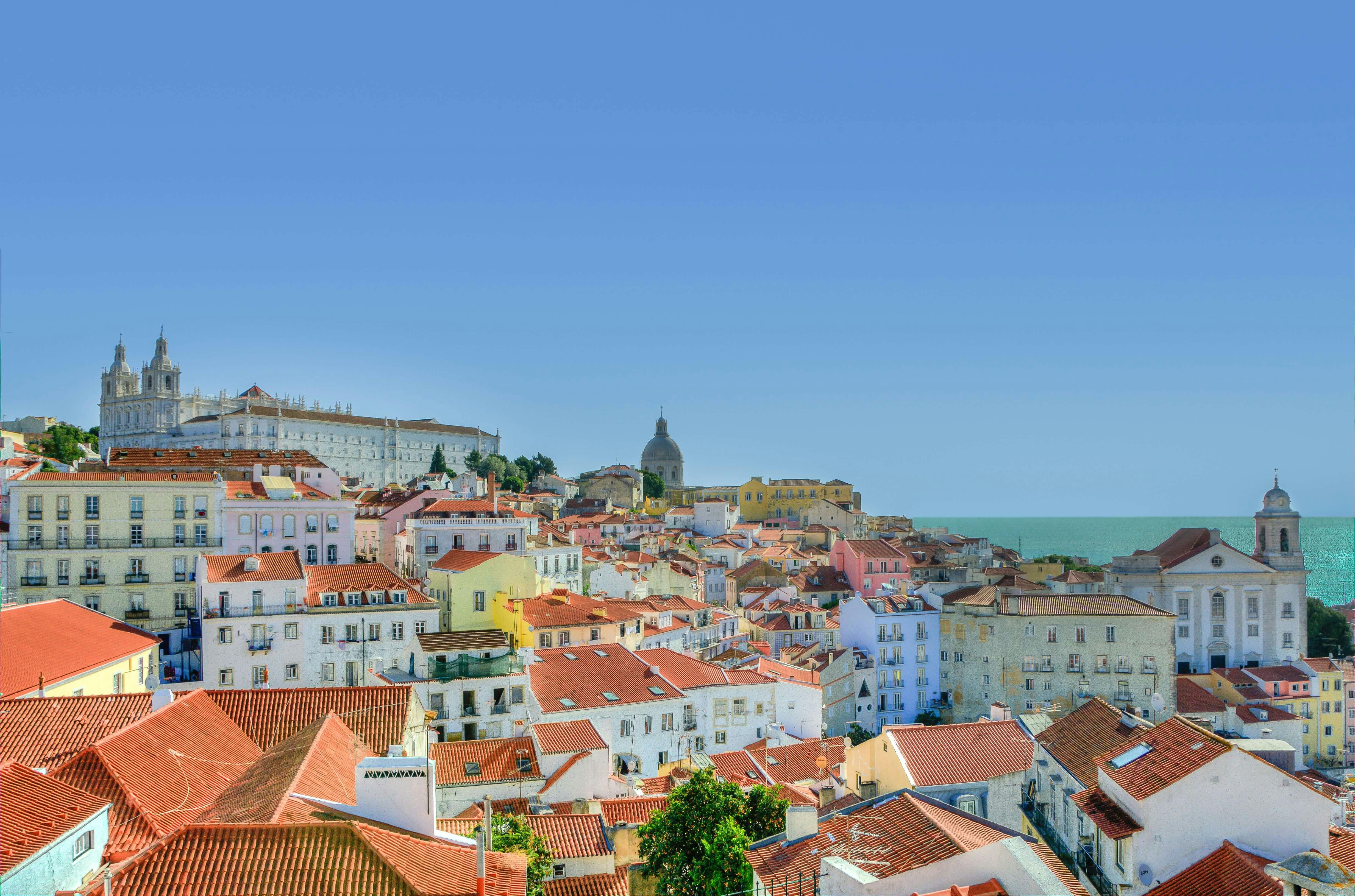 Vista panorâmica de Lisboa a partir do bairro da Sé, mostrando os telhados de terracota da cidade e o Rio Tejo.