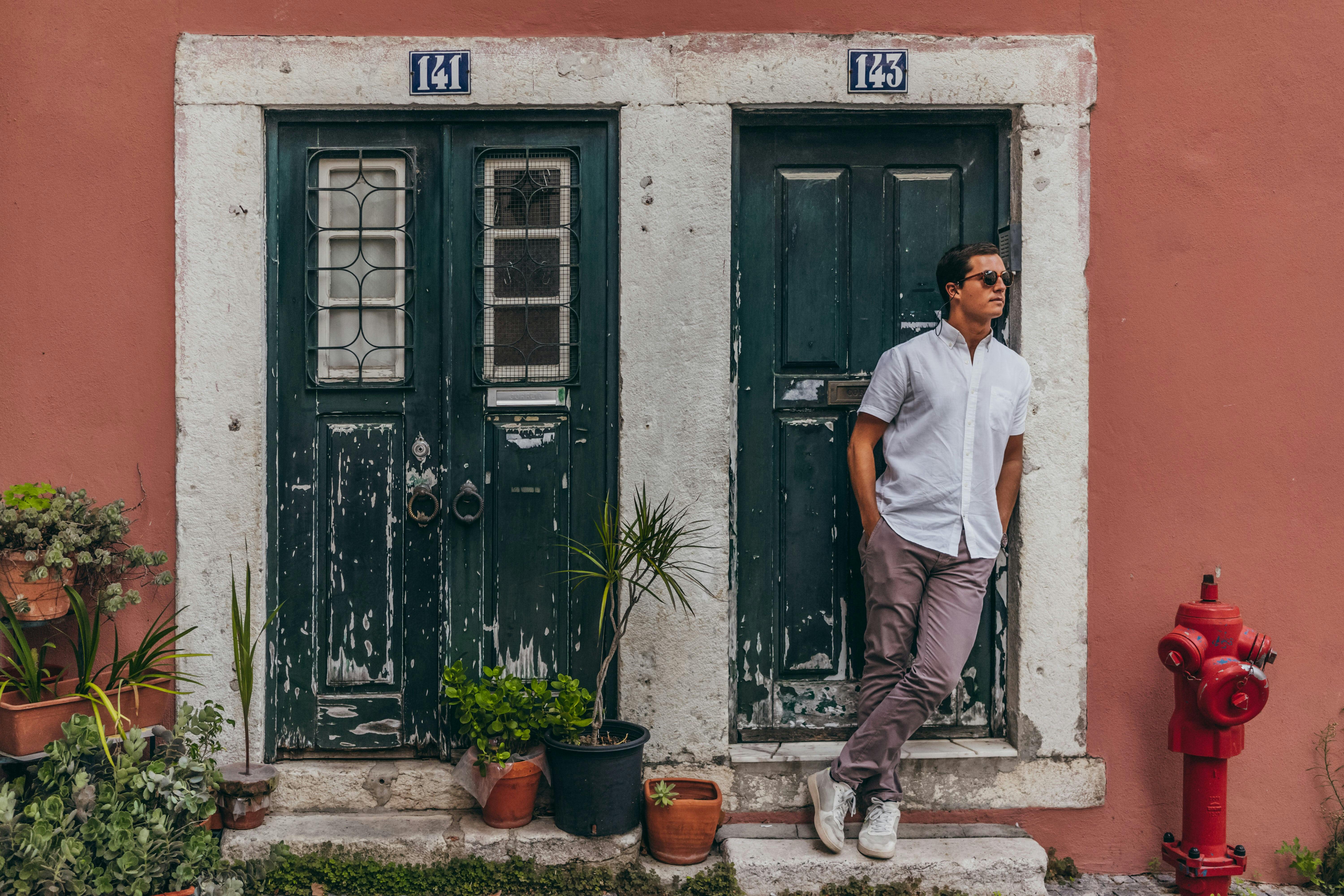 Man with hands in his pockets standing against a vibrant wall featuring two aged doors, numbered 141 and 143, with plants on the doorsteps and a fire hydrant nearby.