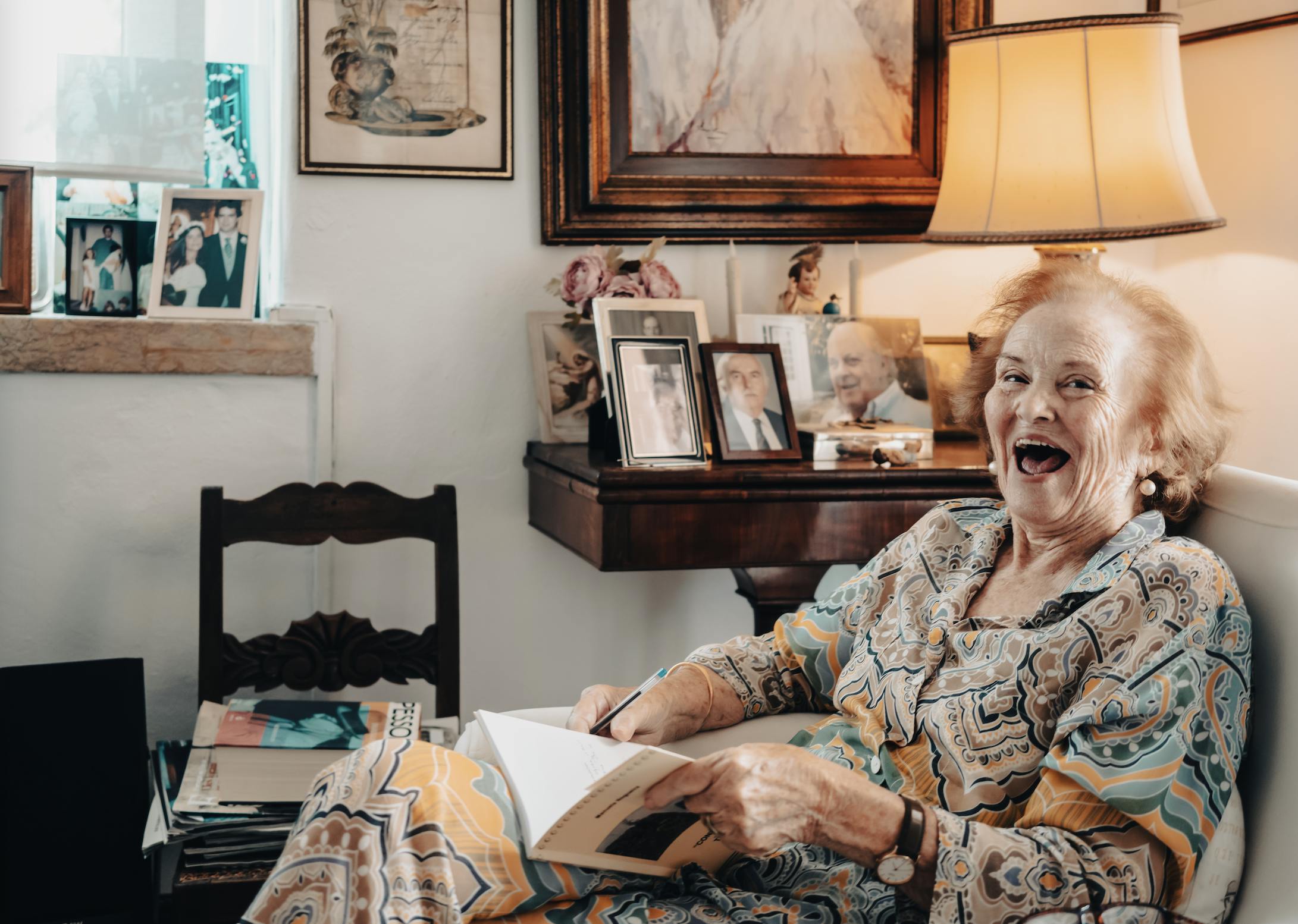 An older woman sitting on her house's white sofa, holding a book and laughing