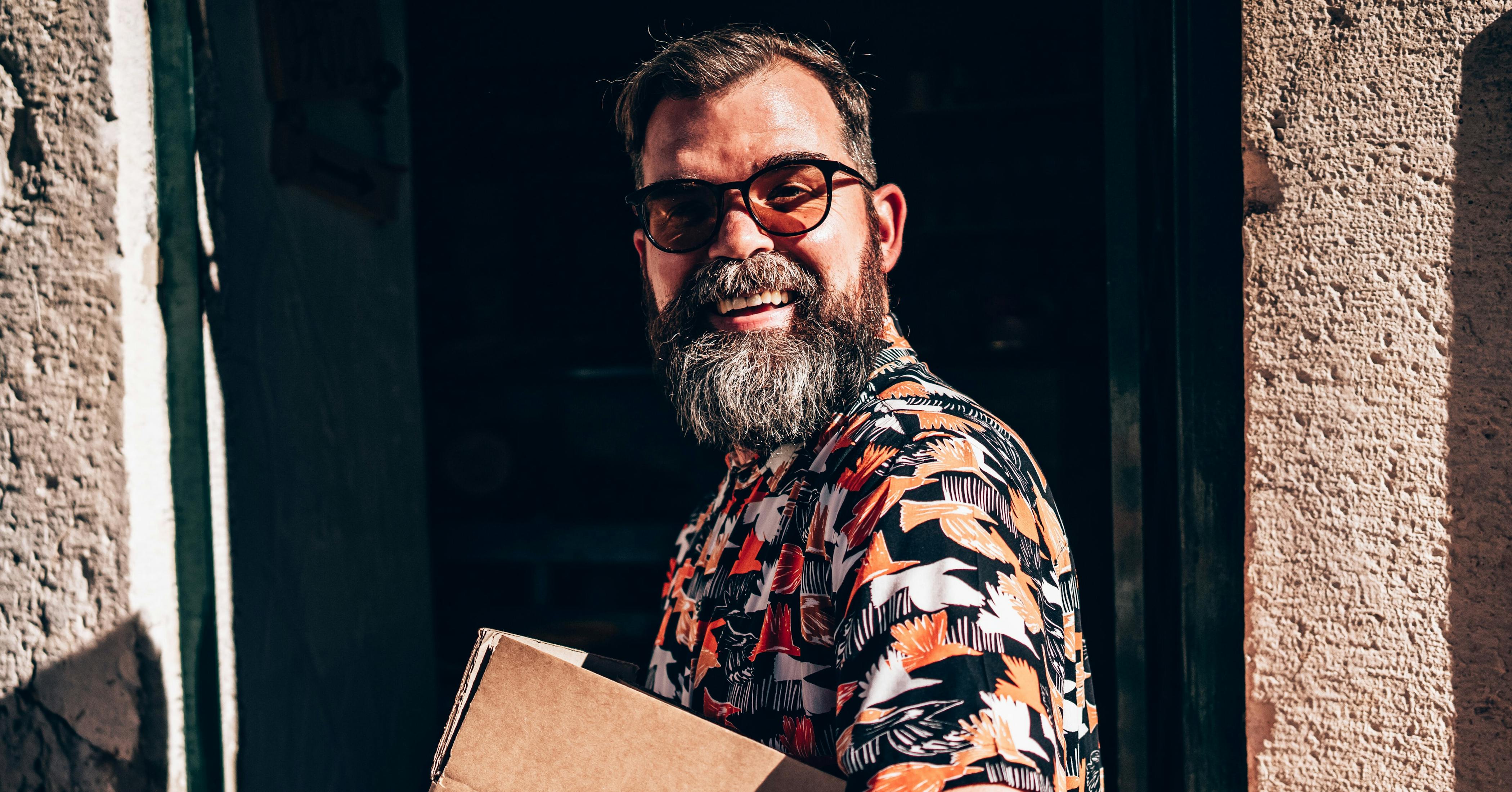 Homem sorridente de óculos escuros e camisa com estampa colorida com temática de natureza, segurando uma caixa de papelão e entrando na porta de um prédio na rua.