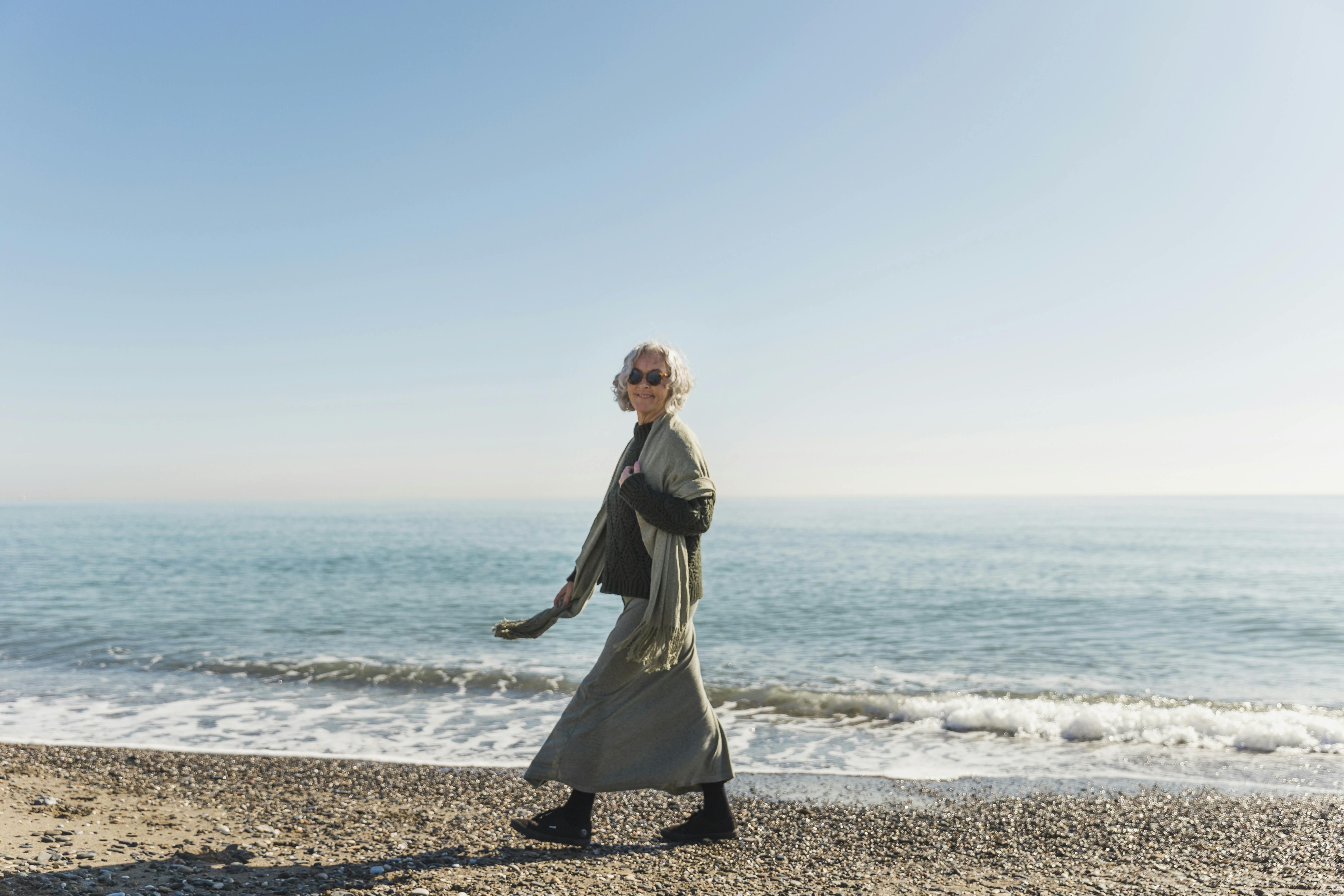 Mulher de cabelos brancos vestida com um xale, caminhando ao longo da costa de uma praia.