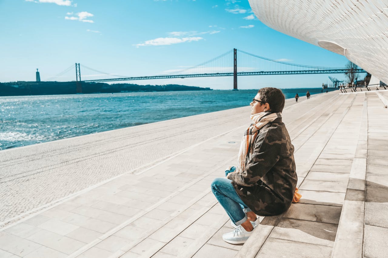 Person sitting on stairs looking at the Lisbon sea view, with Cristo Rei and 25 de Abril bridge on the side