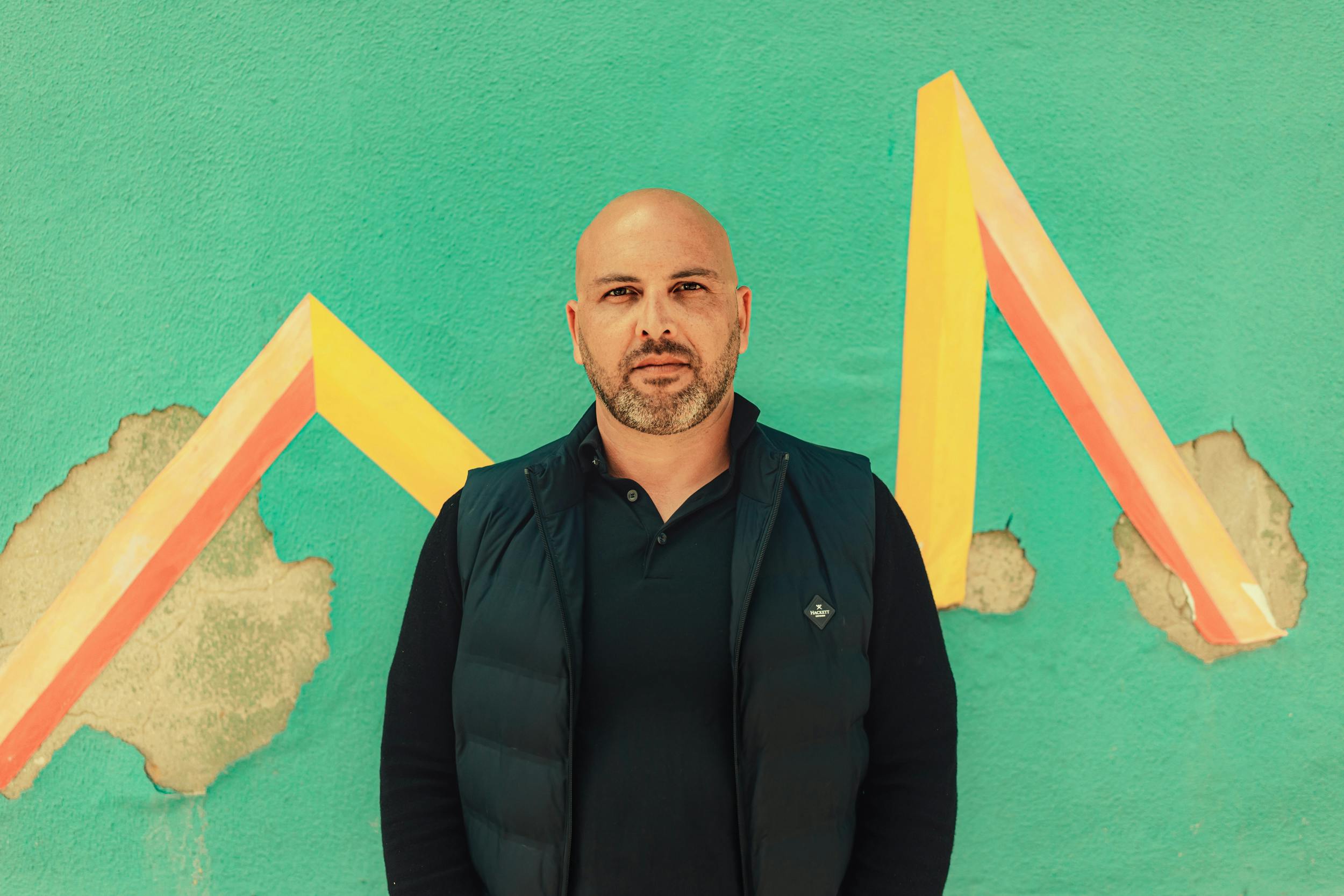 Bald man looking serious in front of a green wall with the letter M drawn