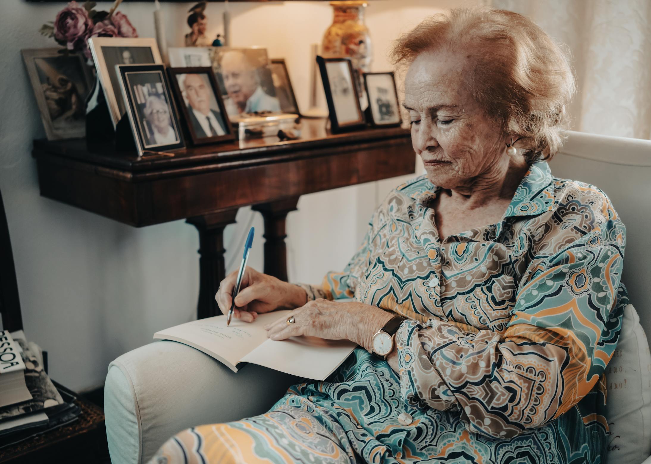 An older woman sitting on her house's white sofa, writing in her notebook with a pen