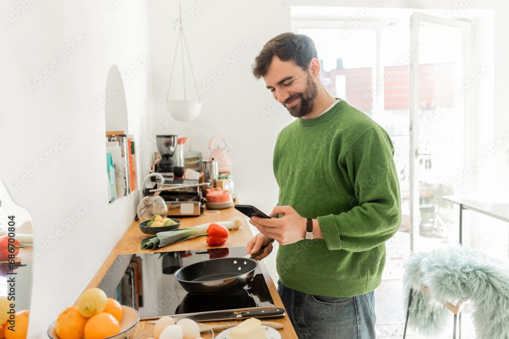 Bearded and brunette digital nomad in casual green jumper looking at the phone and cooking in his kitchen at the same time