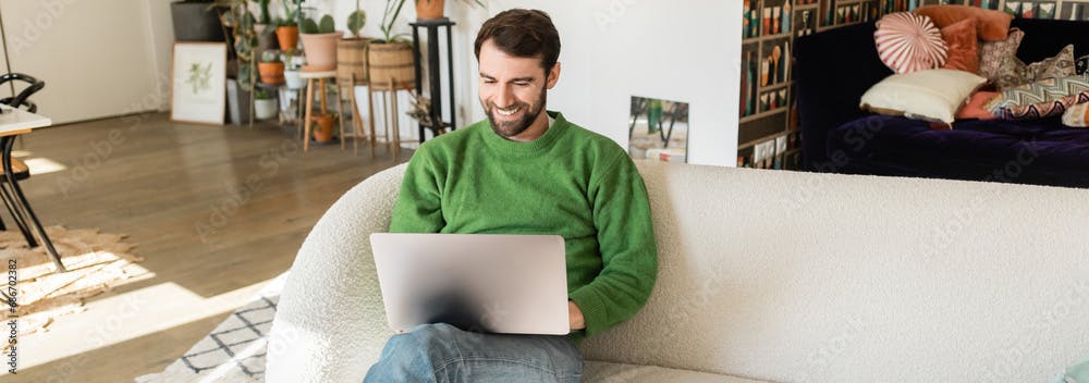 Bearded and brunette digital nomad in casual green jumper sitting on his sofa, on the living room with a laptop on his lap.