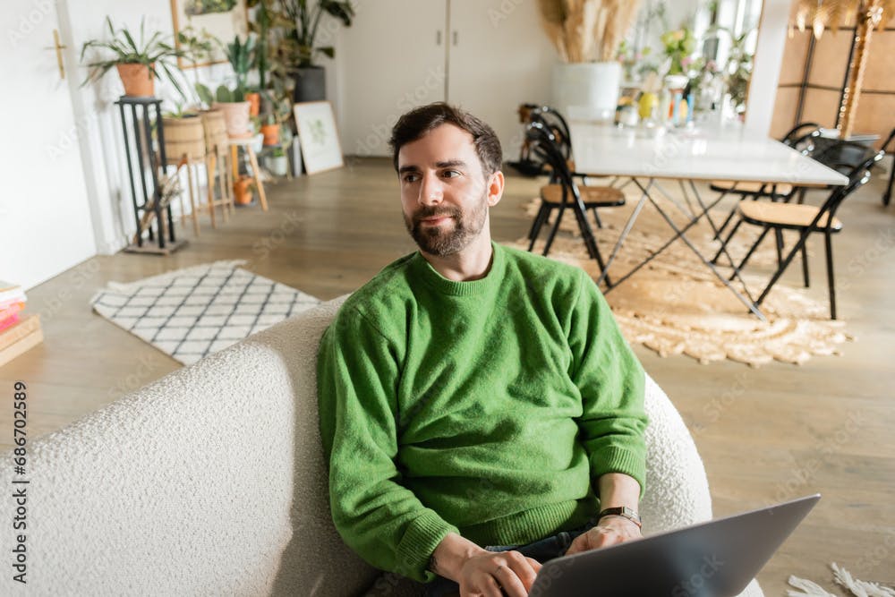 Bearded and brunette digital nomad in casual green jumper looking away while using laptop at home