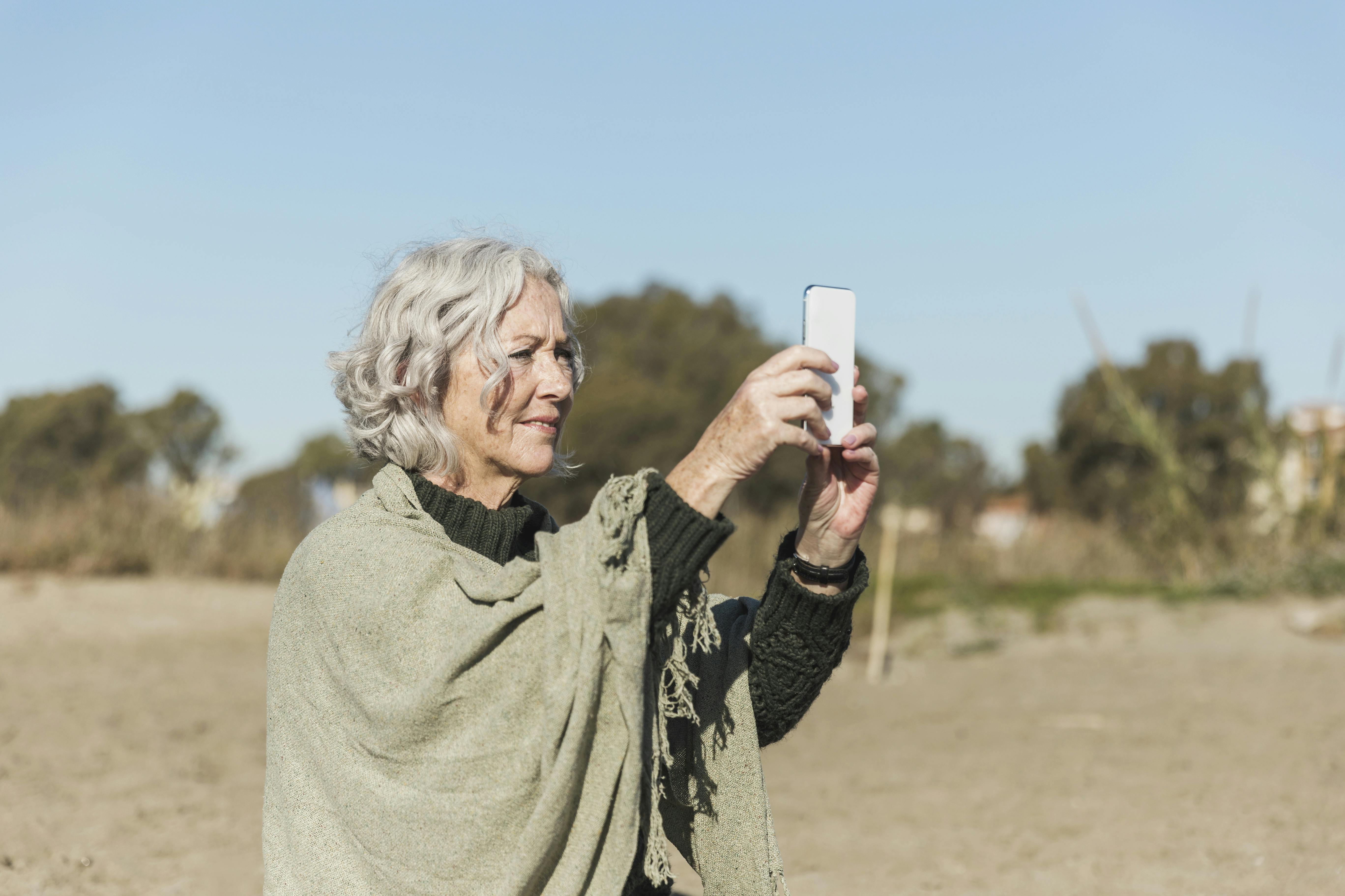 Mulher de cabelos brancos tirando fotos da natureza com seu celular.
