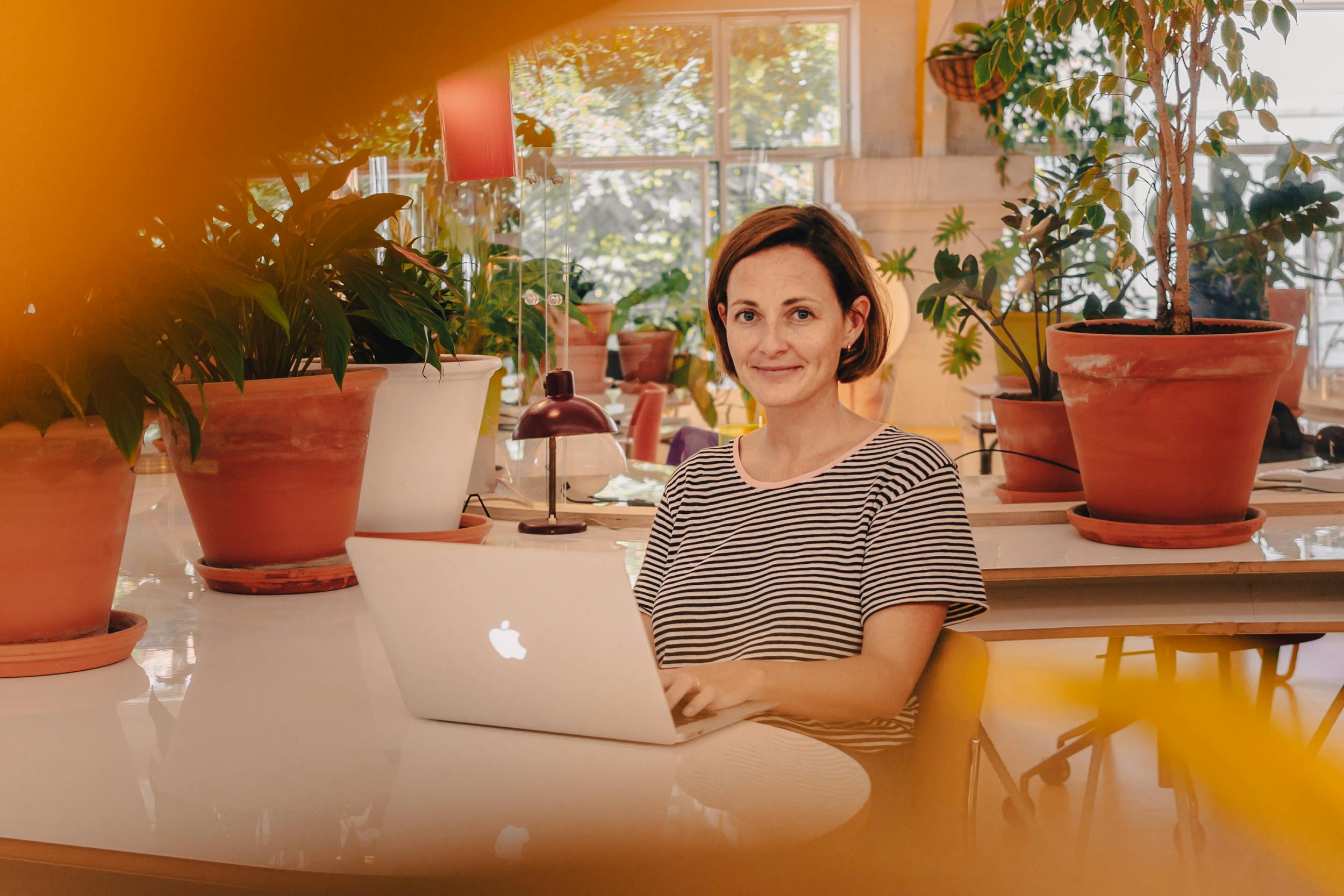 Mulher com cabelo curto sentada em frente a um computador, posando com um sorriso e cercada por vasos de plantas