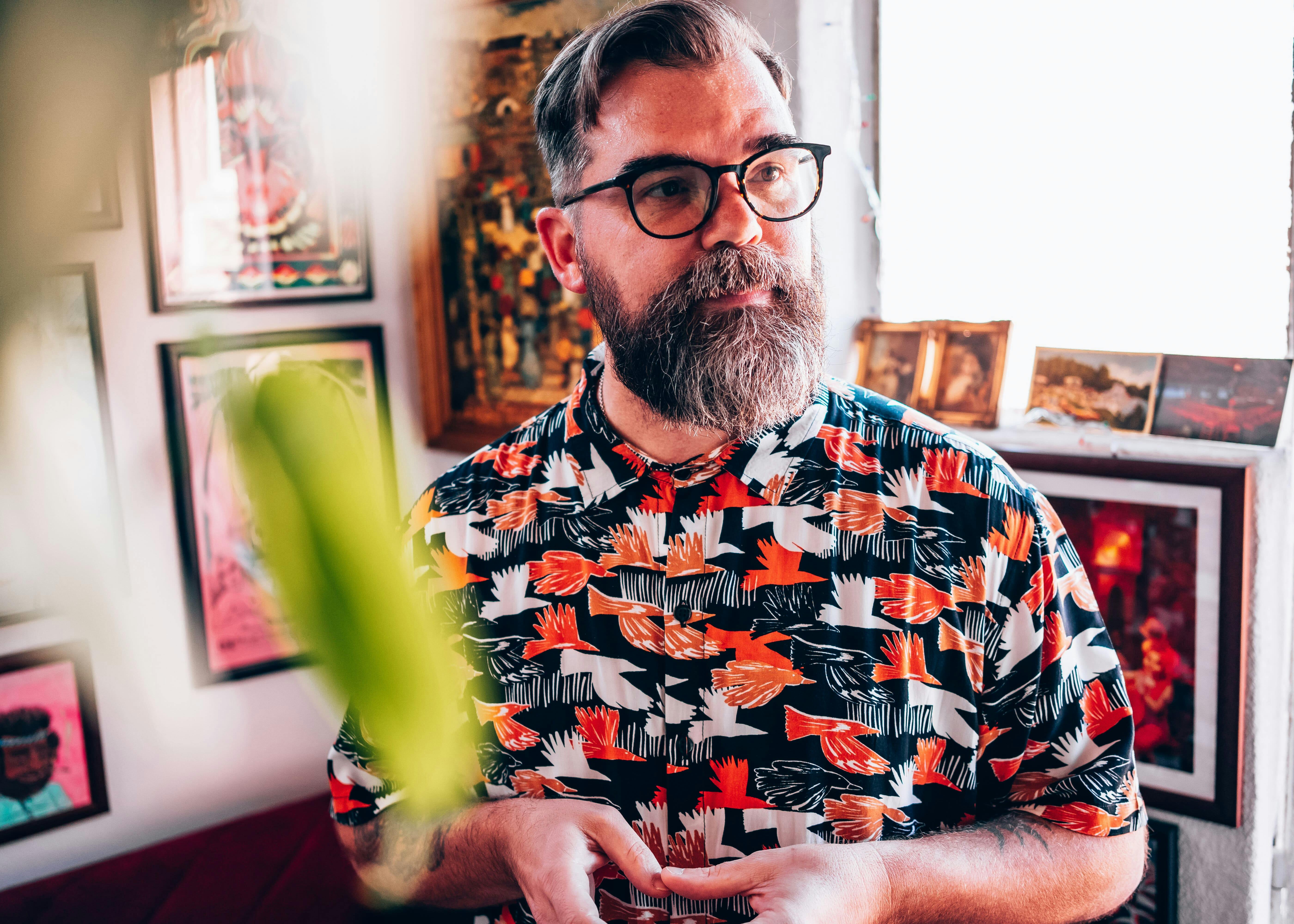 Man in glasses wearing a shirt with a vibrant nature-themed print, looking to the side, with old records displayed on the wall behind him.