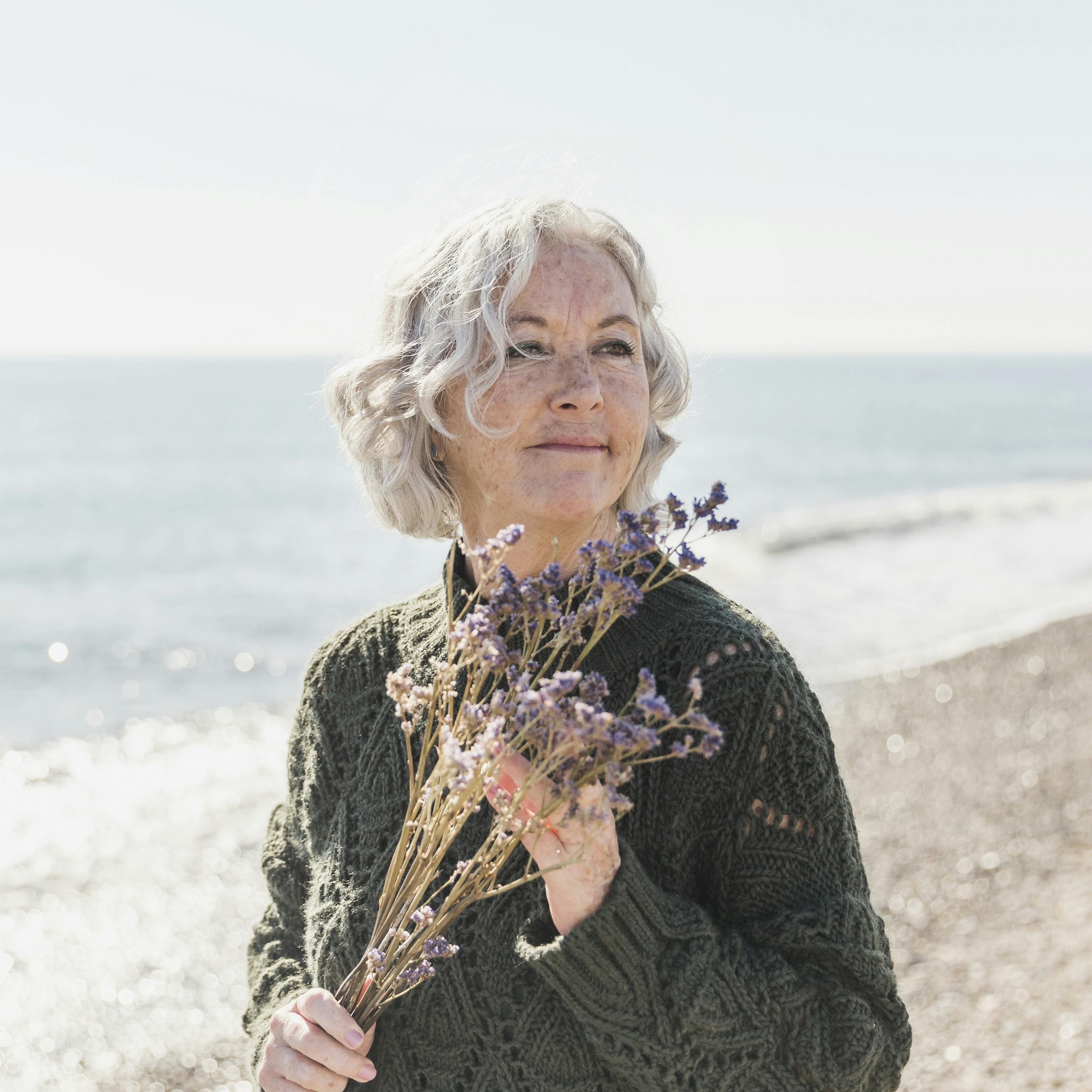 White-haired woman at the beach holding a bouquet of dried flowers.