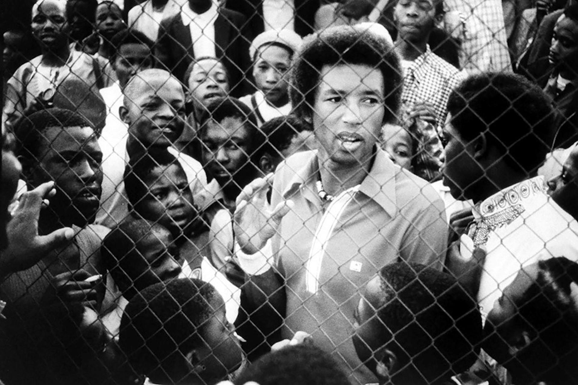 Arthur Ashe in Soweto behind a chain link fence surrounded by a group of young black people.
