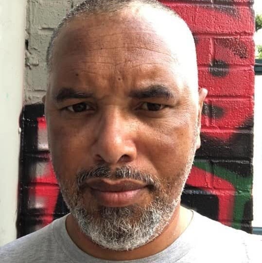 Photo of Sean Jacobs. Sean's skin is medium brown, and he has close cropped salt and pepper hair and a similar beard.