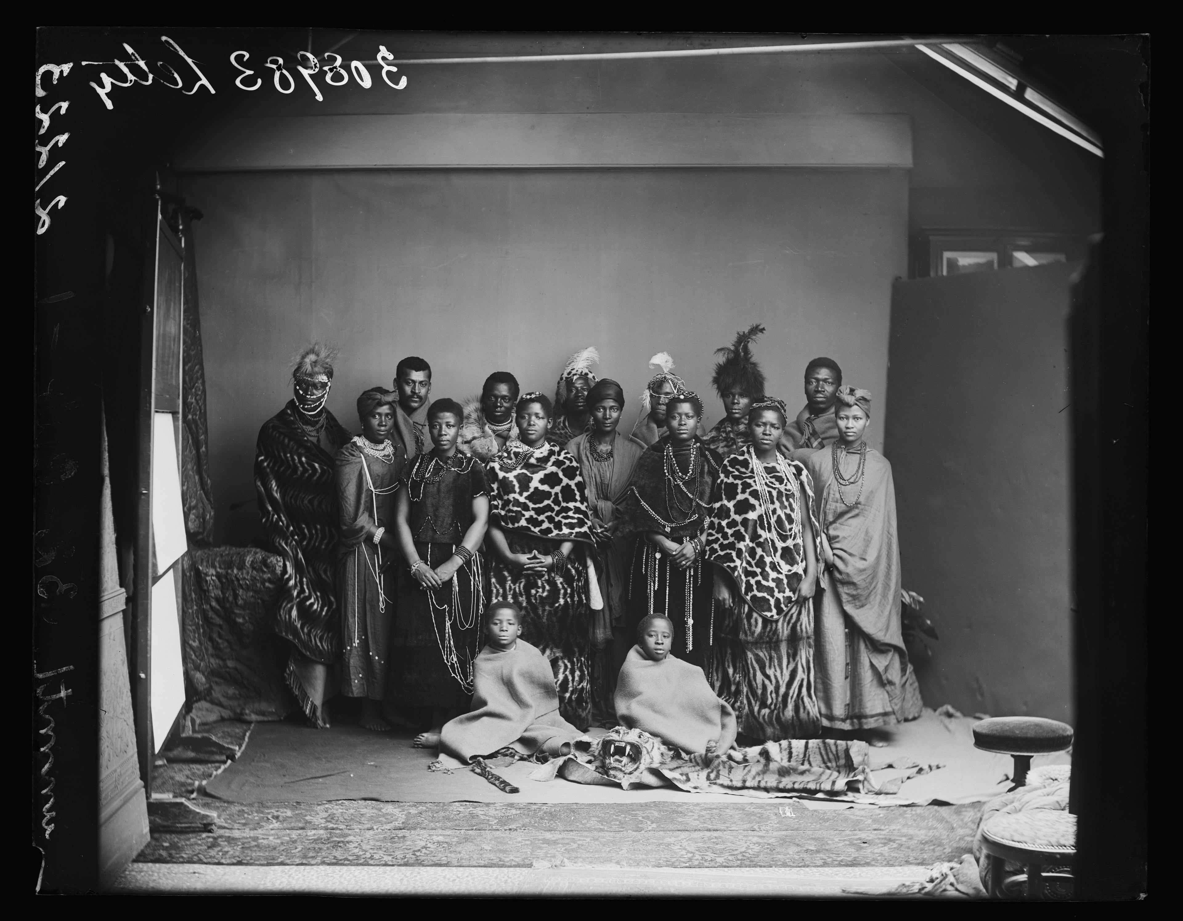 A black and white photo showing a group of Black people in traditional dress. Standing are 14 adults, men and women. Seated at their feet are two children with a tiger skin beneath them.