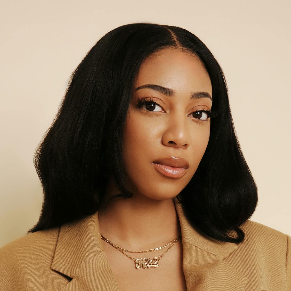 A photo of Mia Coleman. She is a light-skinned Black feminine presenting person, with mid-length hair, worn on in a blown out style with a centre part. She wears a light brown/beige jacket, and a gold double chain necklace with "Mia" etched in a gold pendant at the centre.