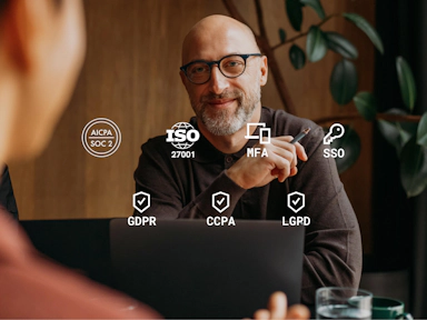 A smiling, bald man with glasses and a trimmed beard sits at a table during a business meeting, holding a pen and facing someone whose back is to the camera. He appears confident and engaged. Overlaid on the image are data privacy and security icons, including AICPA SOC 2, ISO 27001, MFA (multi-factor authentication), SSO (single sign-on), and compliance badges for GDPR, CCPA, and LGPD. The setting is professional with natural light and indoor plants, emphasizing trust, compliance, and secure data practices in a business context.