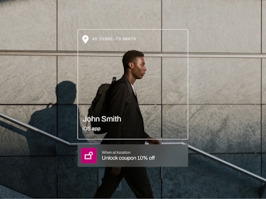 A man in business attire walks past a tiled stone wall while using stairs outdoors, carrying a backpack. Overlaid interface elements display location coordinates (40.70390, -73.98670) and a user profile labeled “John Smith – iOS app.” A promotional banner at the bottom reads, “When at location: Unlock coupon 10% off,” indicating a location-based mobile marketing with a customer data platform.