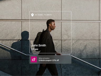 A man in business attire walks past a tiled stone wall while using stairs outdoors, carrying a backpack. Overlaid interface elements display location coordinates (40.70390, -73.98670) and a user profile labeled “John Smith – iOS app.” A promotional banner at the bottom reads, “When at location: Unlock coupon 10% off,” indicating a location-based mobile marketing with a customer data platform.