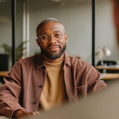 A man with a neatly groomed beard and short-cropped hair sits in a modern office, looking directly at the camera with a calm, confident expression. He wears round glasses, a tan sweatshirt, and a rust-colored corduroy overshirt. The background features soft lighting, glass partitions, a desk lamp, and a hint of greenery, creating a warm, professional atmosphere that suggests trust and clarity.