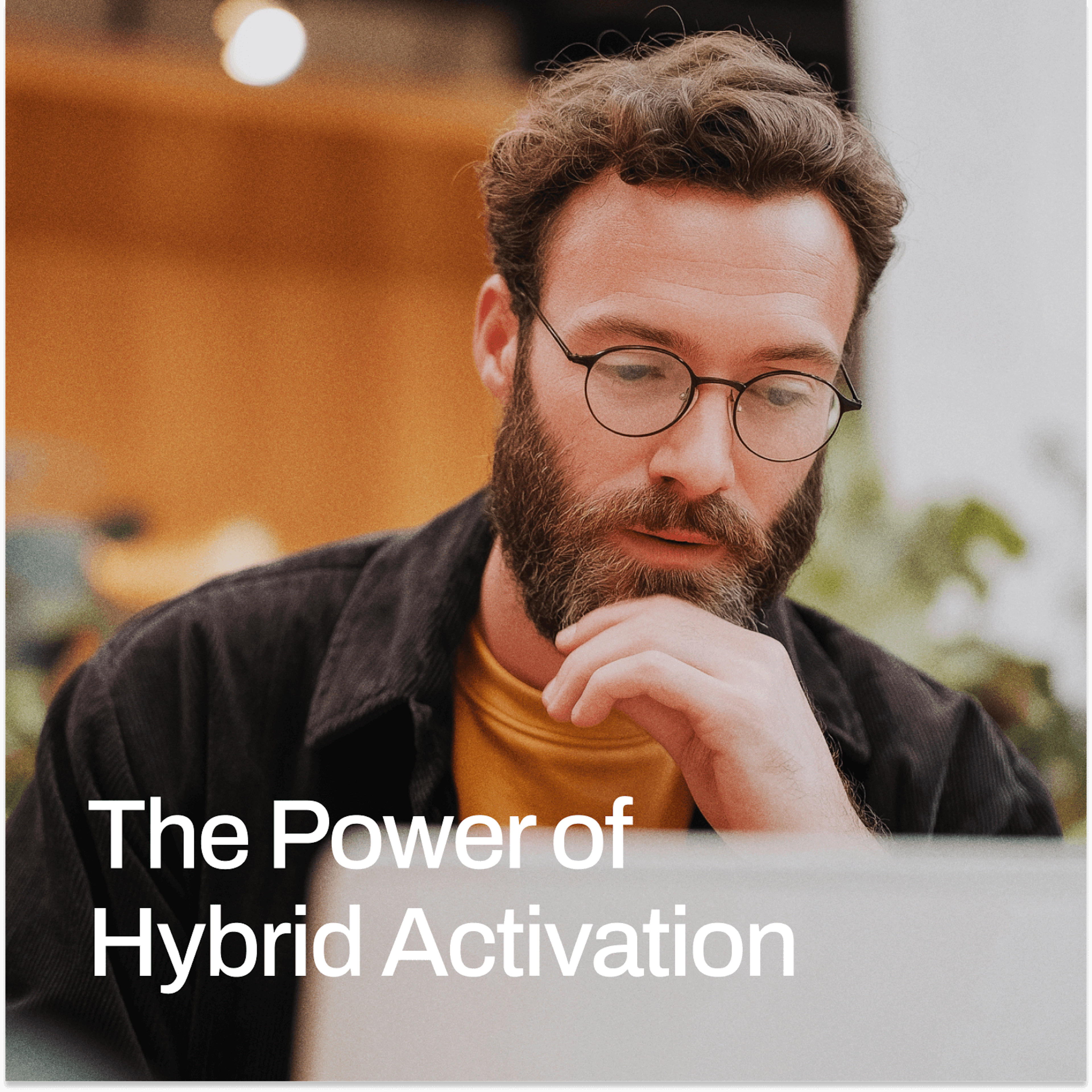 A bearded man with glasses sits thoughtfully in front of a laptop, resting his chin on his hand as he focuses on the screen. He wears a mustard yellow shirt under a dark overshirt. The background is softly blurred with warm lighting and indoor plants, suggesting a modern co-working space or office. White text in the foreground reads, “The Power of Hybrid Activation.”