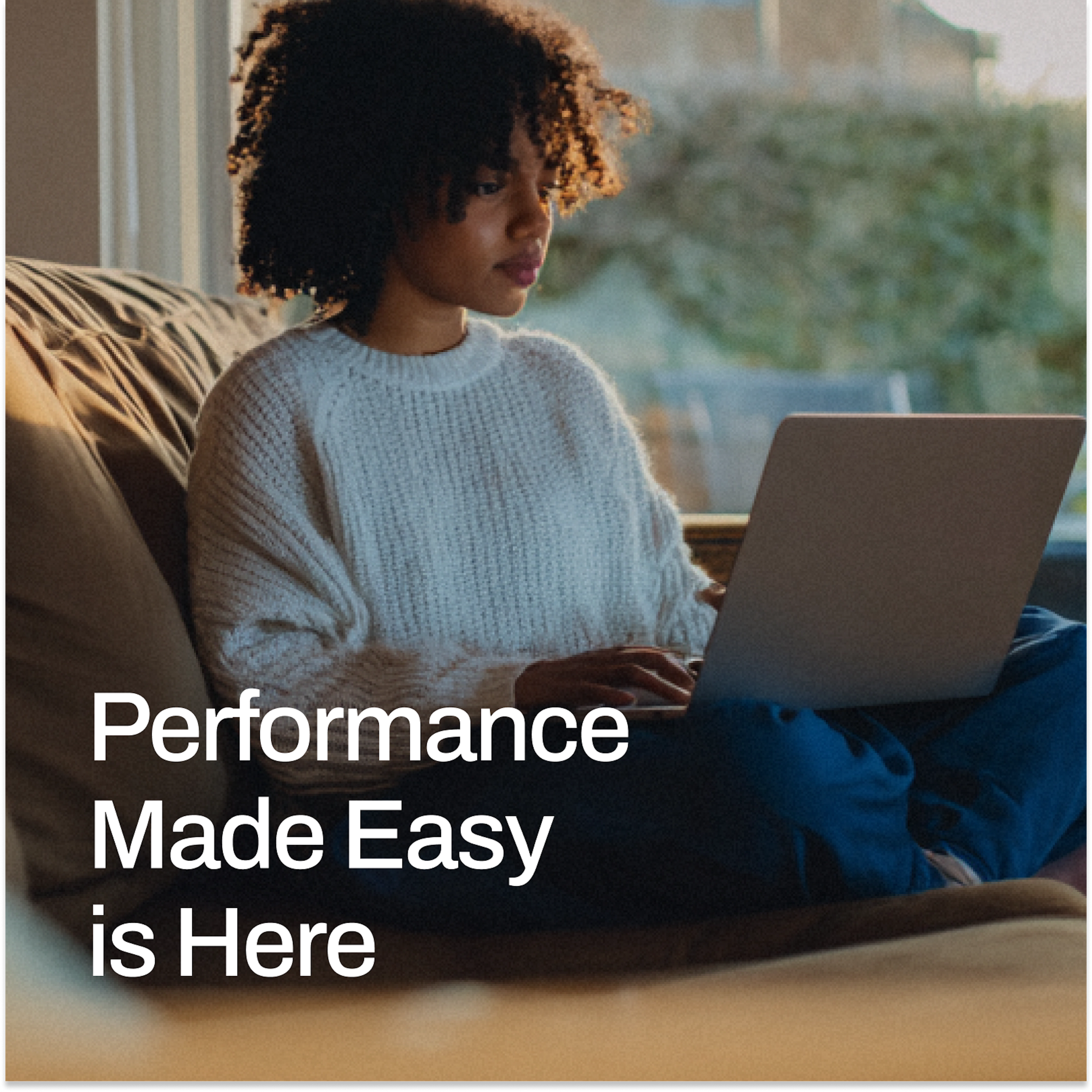 A young woman with curly hair types on a laptop while sitting on a couch by a sunlit window, with bold white text reading “Performance Made Easy is Here.”