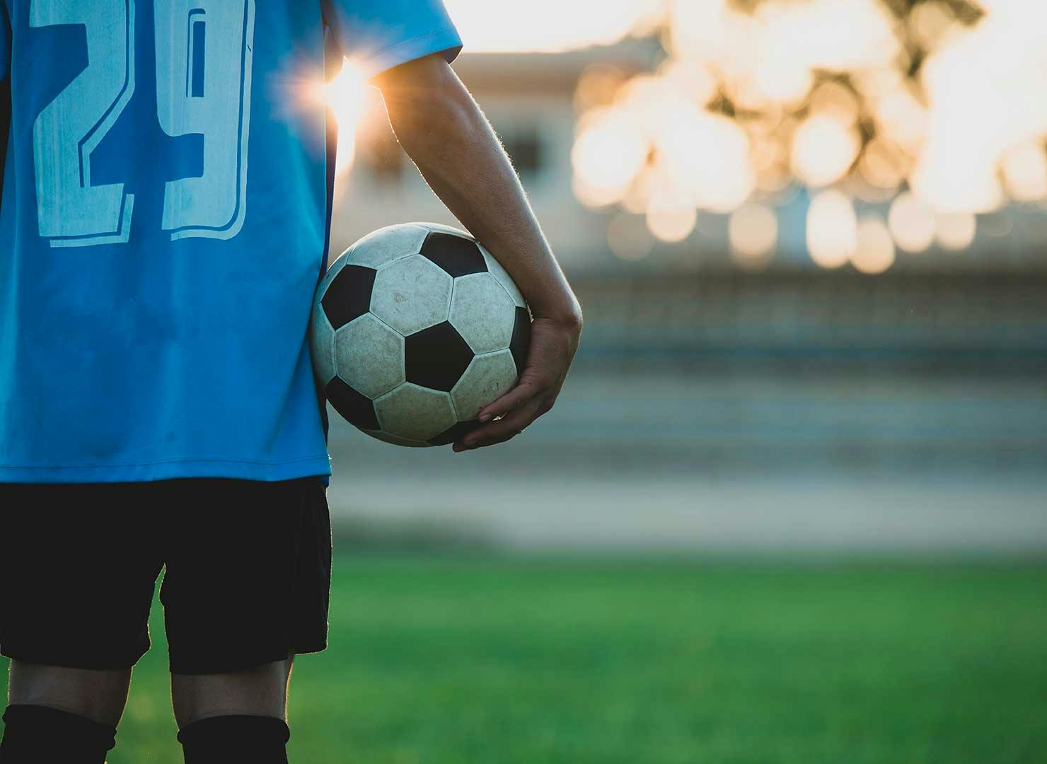 A soccer player in a blue jersey with the number 29 holds a soccer ball under their arm on the field, with the sun setting in the blurred background.