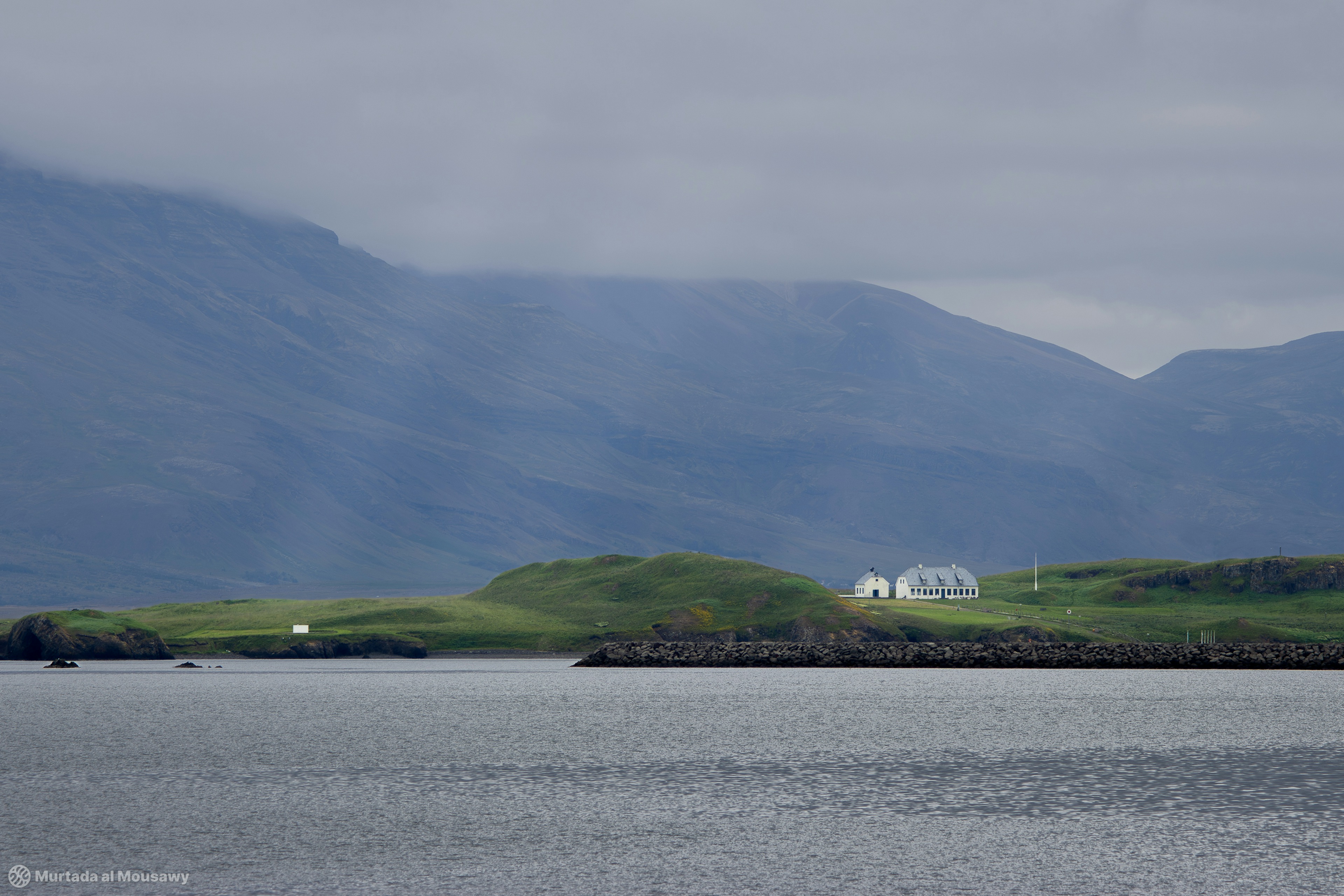 Photo of a distant peninsula with a white building set against large mountains that look blue because of atmospheric perspective.