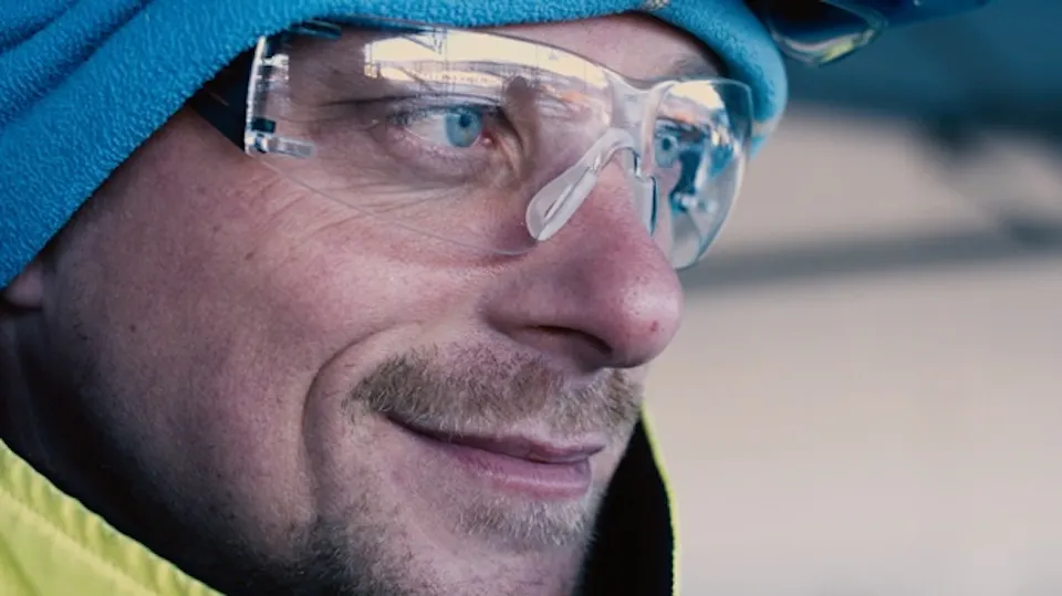 Close-up of a man in clear safety glasses and a blue beanie, smiling, wearing a yellow high-visibility jacket outdoors.