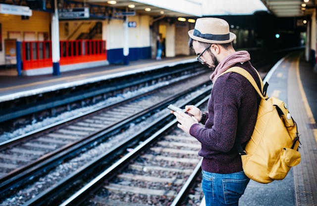 A man with a hat, glasses and a yellow backpack looks at his smartphone while waiting on the platform at a train station.