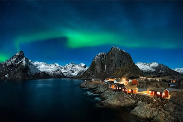 Green northern lights over a coastal landscape with red, illuminated rorbu cabins along the shore and snow-capped peaks behind.