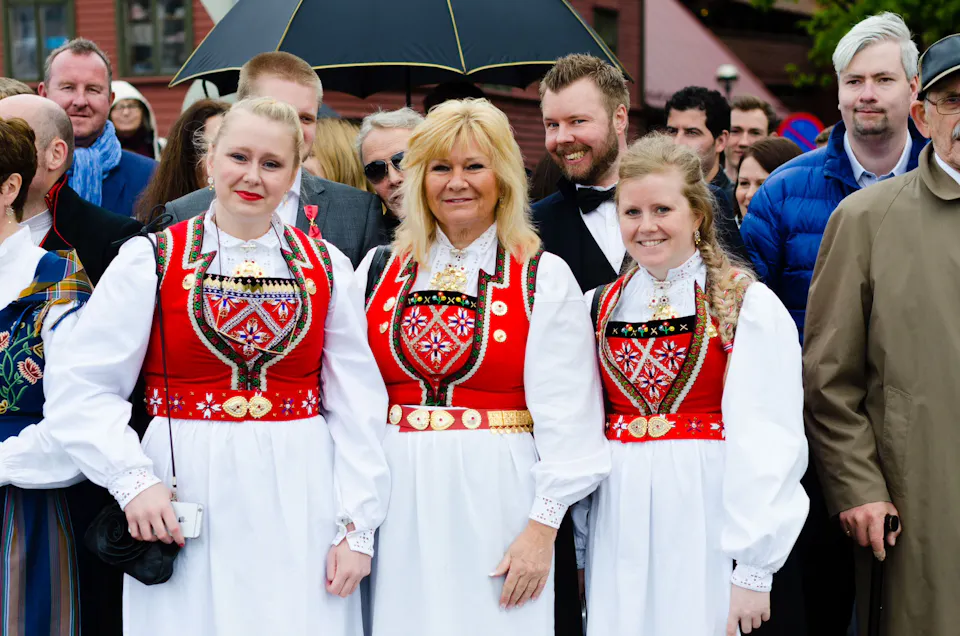 Three women wearing traditional Norwegian bunad costumes with red embroidered bodices and white blouses standing in a crowd outdoors.