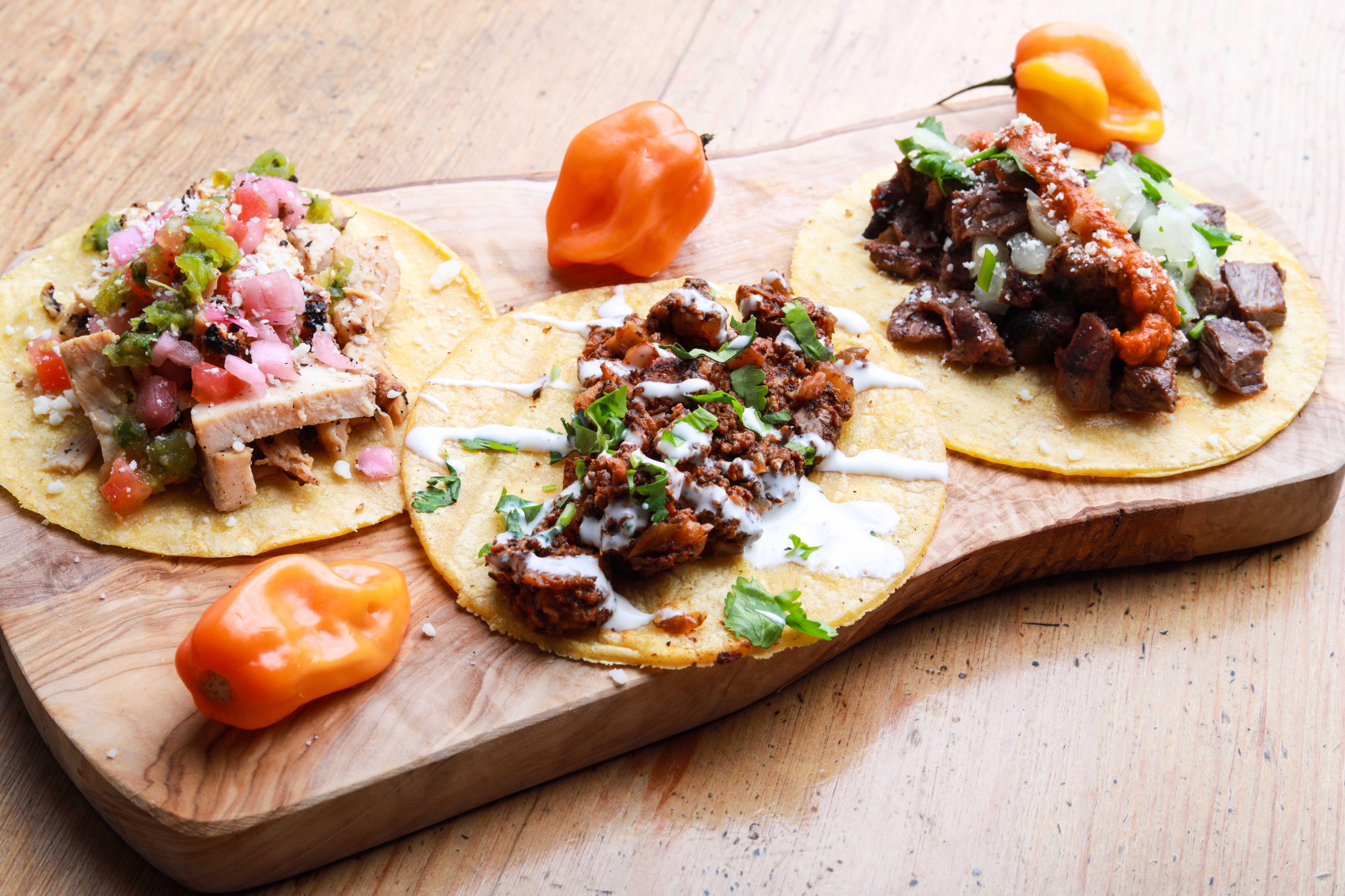 Trzy tacos z tortilli kukurydzianej z różnymi nadzieniami na desce do serwowania, udekorowane kolendrą i pomarańczowym chili.