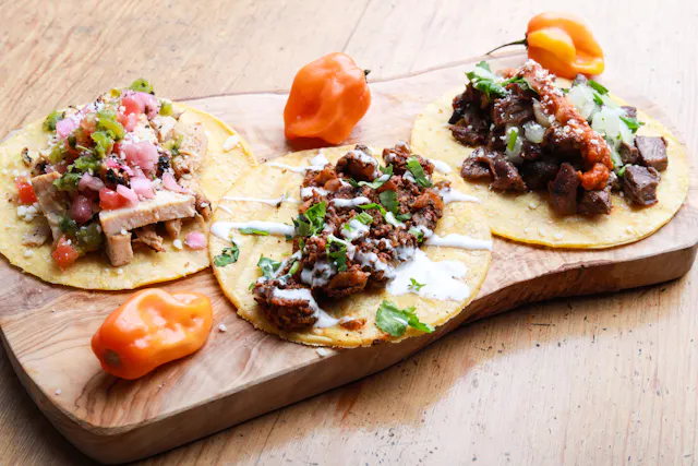 Trzy tacos z tortilli kukurydzianej z różnymi nadzieniami na desce do serwowania, udekorowane kolendrą i pomarańczowym chili.