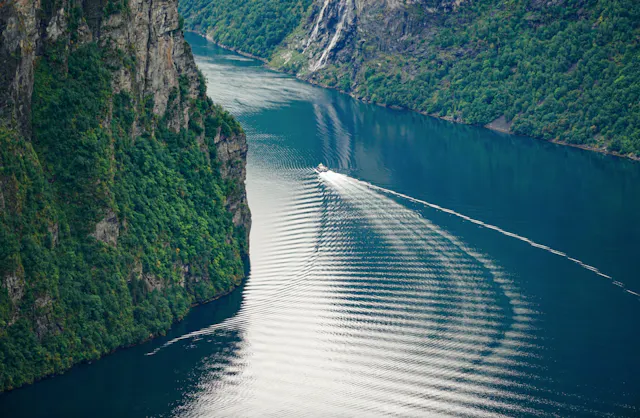 Aerial photo of a narrow fjord with steep green mountain sides and a small boat leaving a wake on the water.