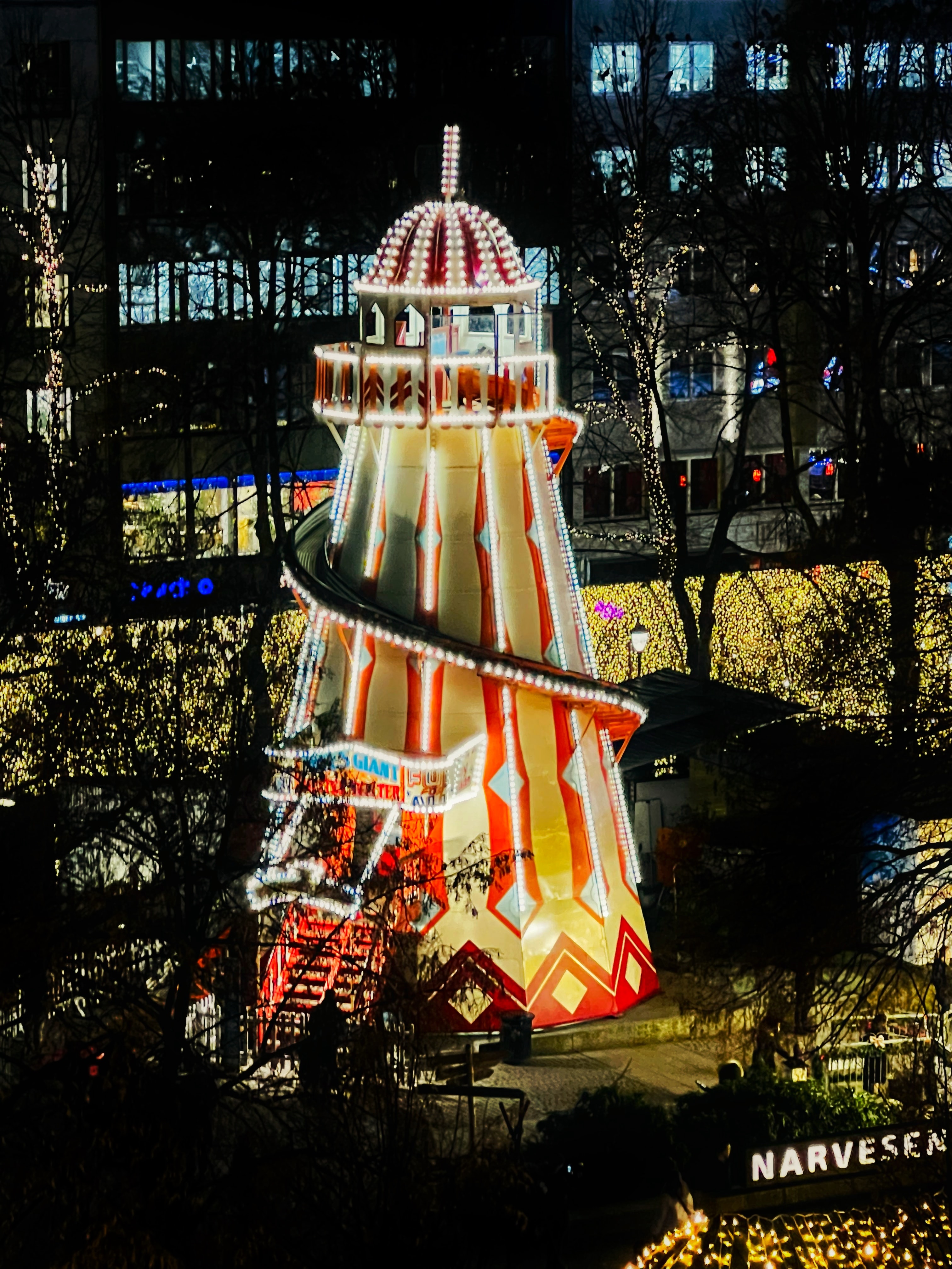 Iluminowana wieża rozrywkowa ze spiralną zjeżdżalnią ozdobiona czerwonymi i białymi światłami nocą z szyldem Narvesen poniżej.