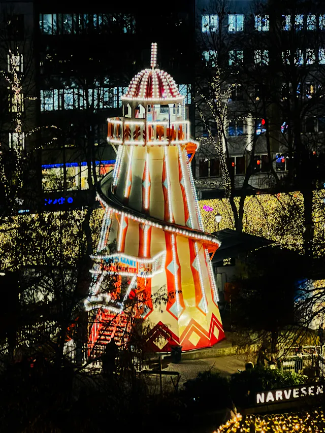Iluminowana wieża rozrywkowa ze spiralną zjeżdżalnią ozdobiona czerwonymi i białymi światłami nocą z szyldem Narvesen poniżej.