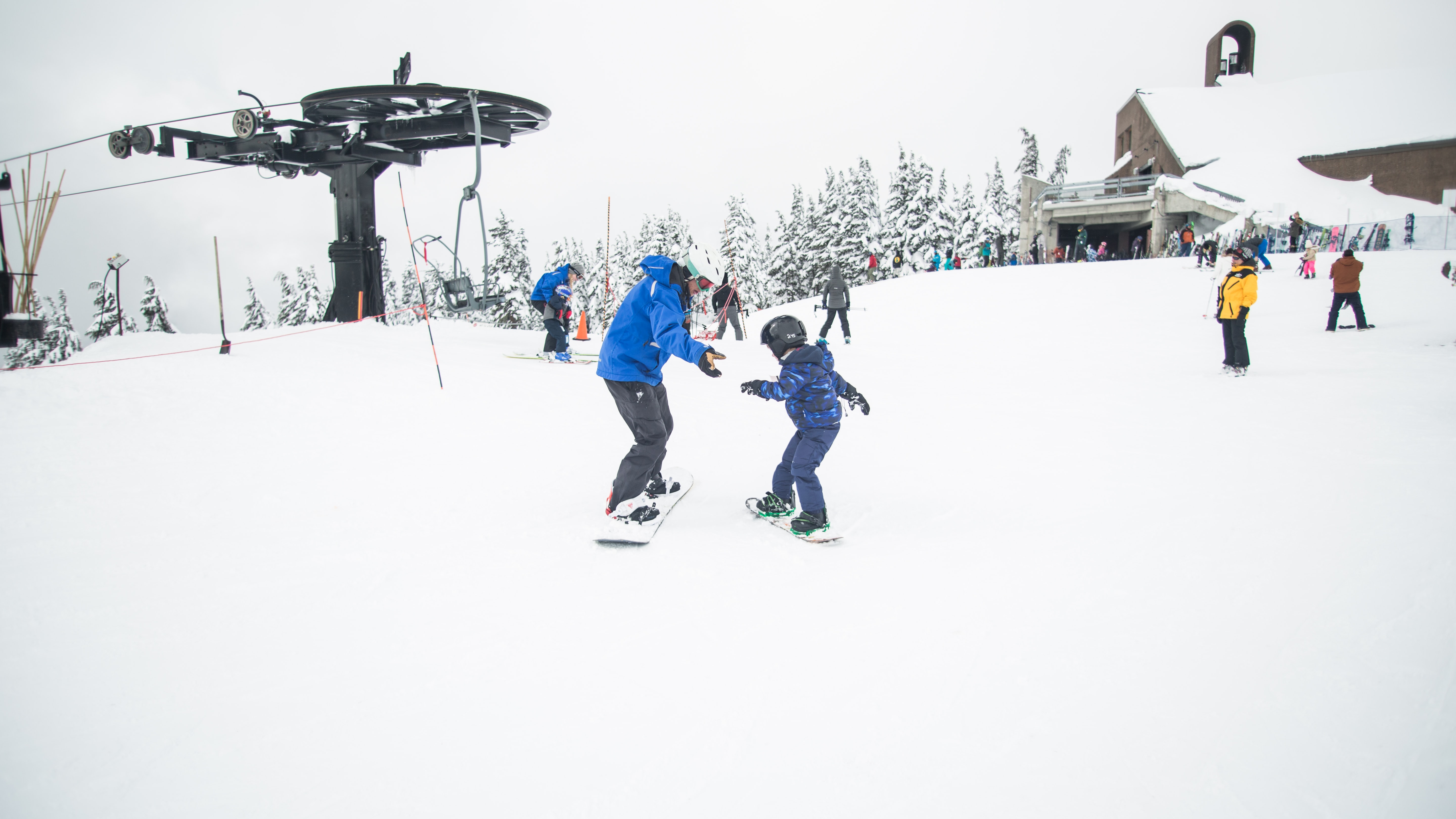 Dorosły w niebieskiej kurtce uczący dziecko jazdy na snowboardzie na ośnieżonym stoku z wyciągiem narciarskim w tle.