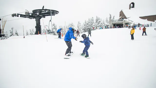 Dorosły w niebieskiej kurtce uczący dziecko jazdy na snowboardzie na ośnieżonym stoku z wyciągiem narciarskim w tle.