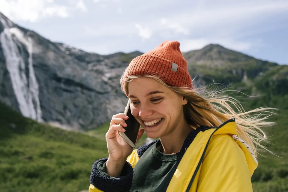 Uśmiechnięta młoda kobieta w czerwonej czapce i żółtej kurtce rozmawia przez telefon na tle norweskich gór i wodospadu.