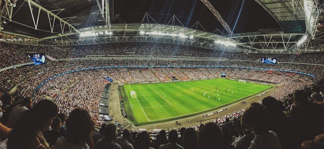 Panoramic view of a large football stadium at night filled with spectators and a bright green pitch illuminated by floodlights.