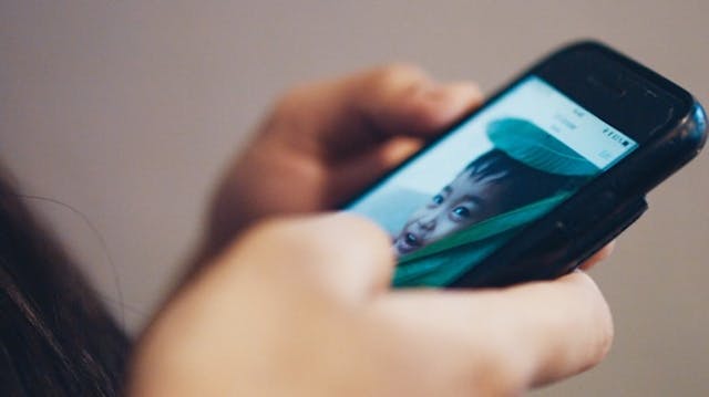 Ręka trzyma ciemny smartfon pokazujący zbliżenie rozmowy wideo z młodą osobą o zaskoczonym wyrazie twarzy.