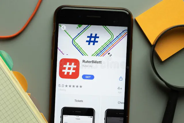 Smartfon leżący na biurku wyświetla aplikację RuterBillett w sklepie z aplikacjami, obok widoczny notatnik i lupa.