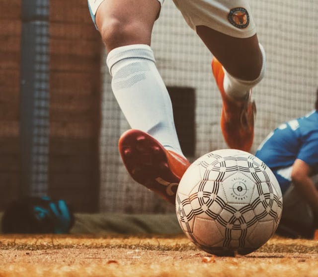 Close-up of a football boot about to kick a ball on sandy ground, with a player in a blue kit in the background.