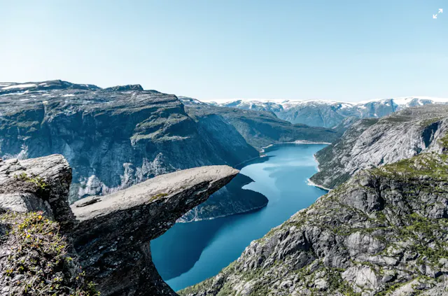 The Trolltunga formation juts out over a deep blue fjord with steep mountain walls and snow-capped peaks in the background.
