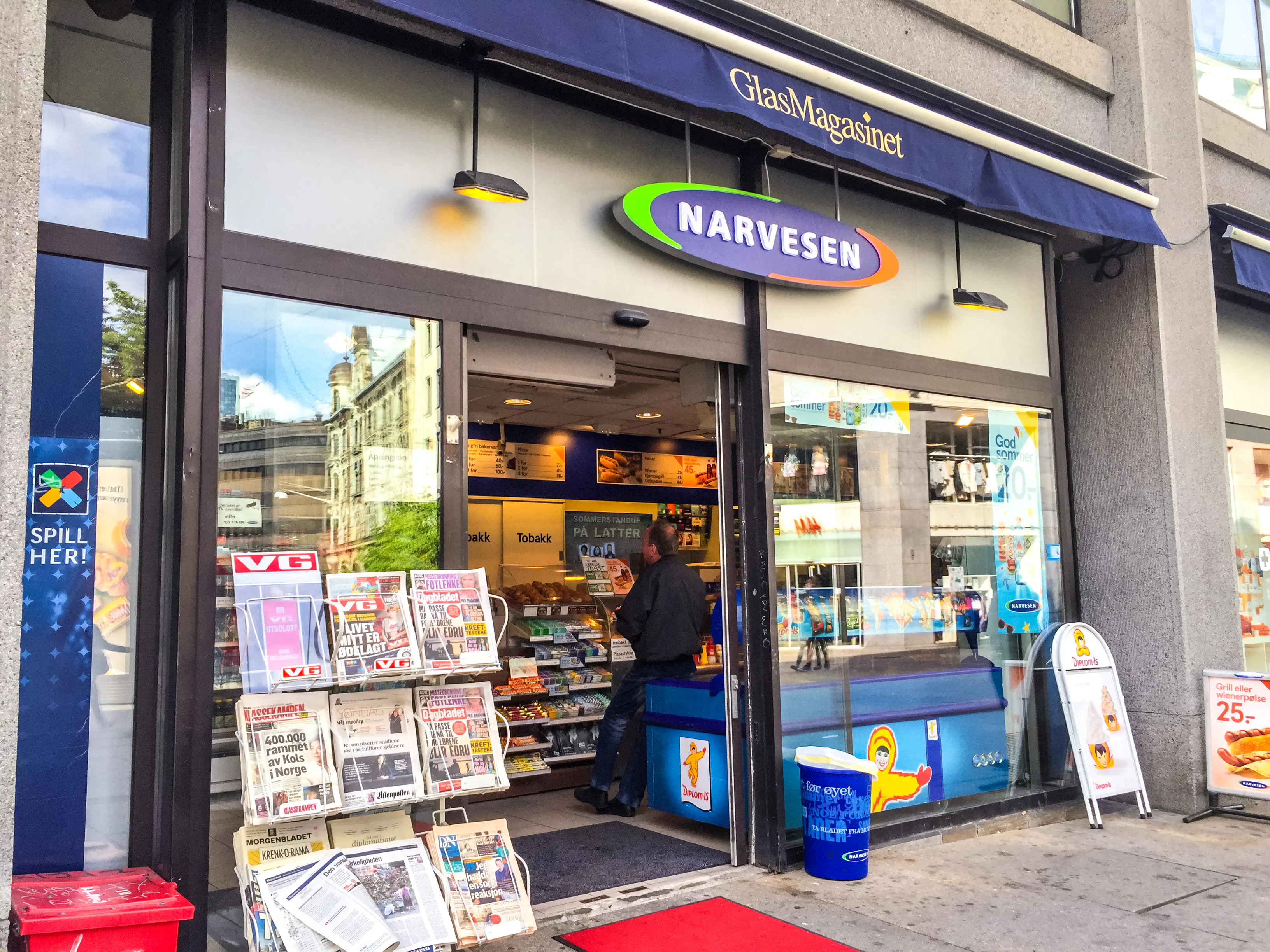 A Narvesen convenience store in Norway with newspaper displays at the entrance and a person visible inside the shop.