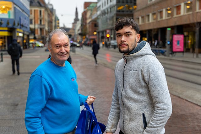 Two men on a city street, an older man in a blue sweater holding blue shopping bags with a younger man in a grey hoodie.