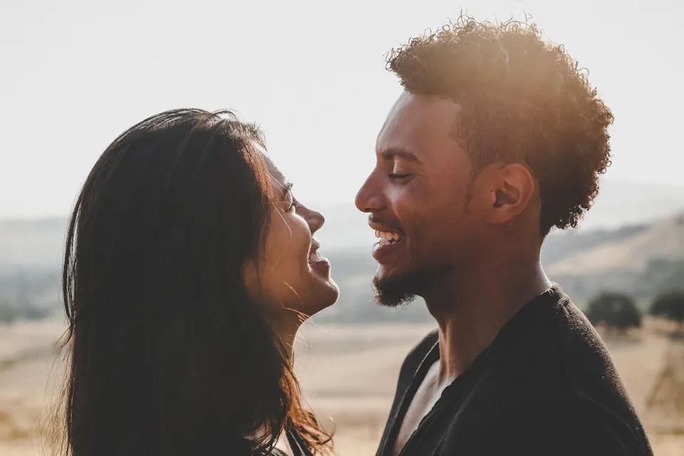 Two people facing each other and laughing outdoors in warm golden sunlight, with rolling hills visible in the background.