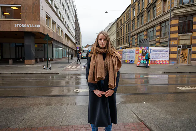 Młoda kobieta w ciemnym płaszczu z brązowym szalem sfotografowana na mokrej od deszczu Storgata z kamienicami w tle.