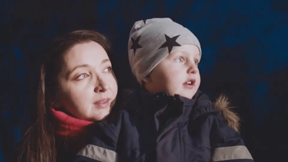 Woman and a young child in winter jackets and a grey star-patterned beanie looking into the distance against a dark background.